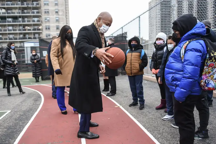 Mayor Eric ADams in a coat and mask holds a basketball in the yard, with school children watching